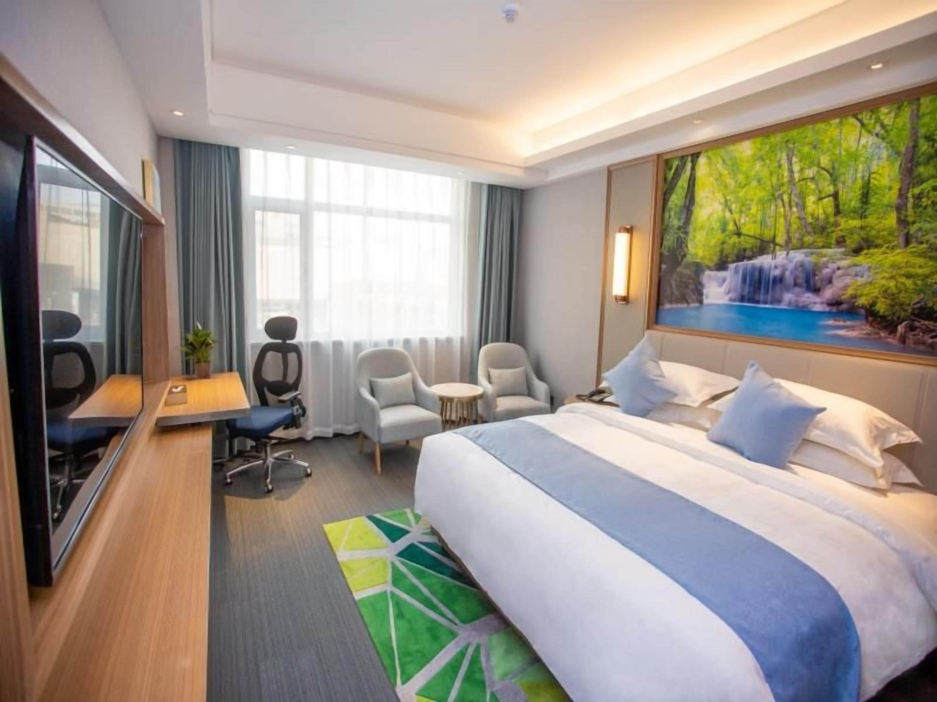A hotel room with a large bed, a desk, two chairs, a mirror, and a painting of a waterfall.