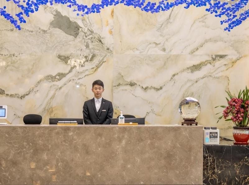 Mostrador de recepción de hotel con pared de mármol, una recepcionista y una planta en maceta.