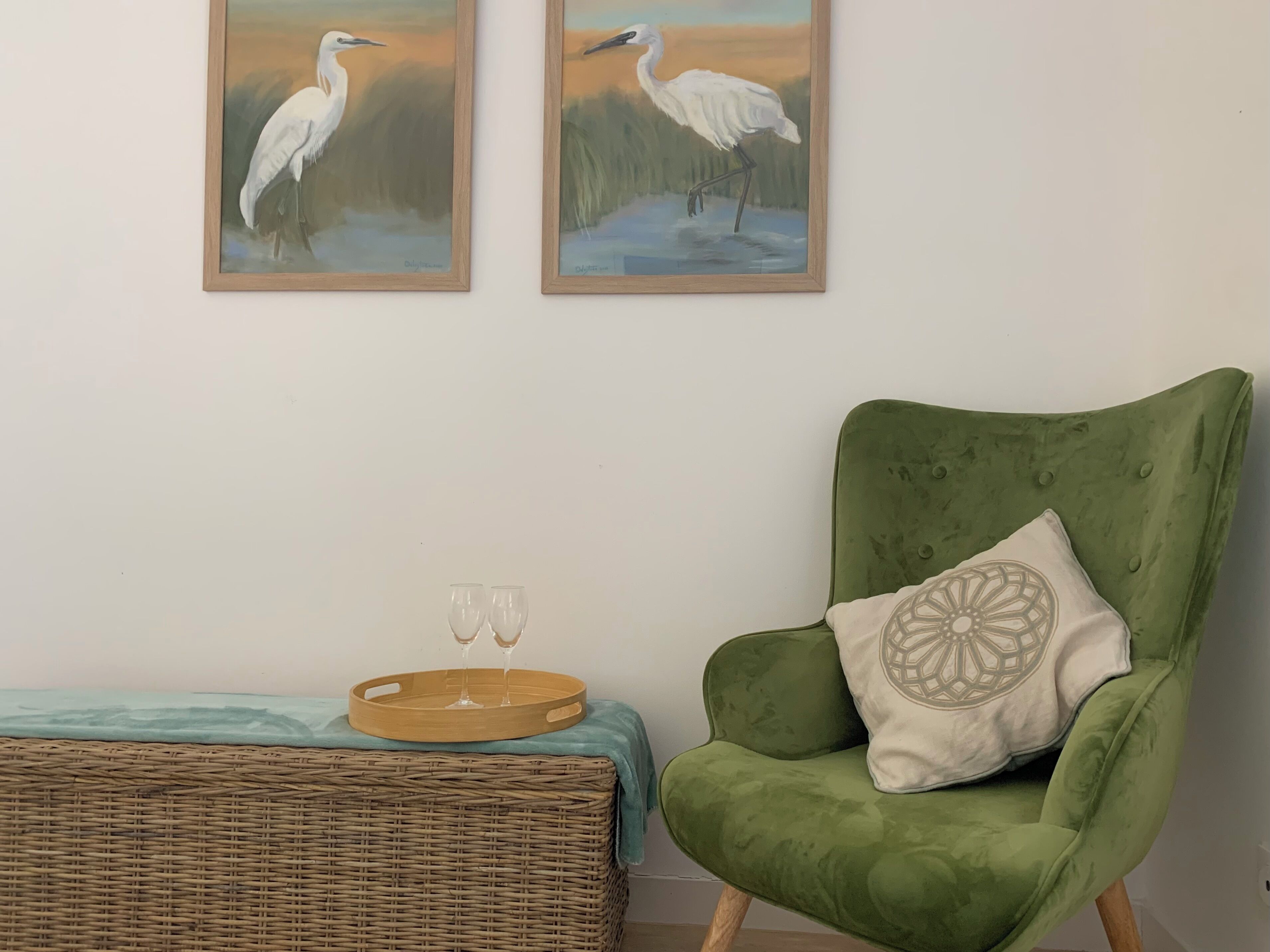 Une pièce comprenant un fauteuil vert, deux tableaux encadrés représentant des oiseaux, une petite table d’appoint en osier avec deux verres et un plateau.
