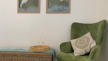 Une pièce comprenant un fauteuil vert, deux tableaux encadrés représentant des oiseaux, une petite table d’appoint en osier avec deux verres et un plateau.