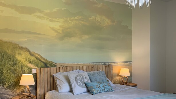 Un dormitorio con una cama grande, dos mesitas de noche con lámparas, una manta azul y un mural con una escena de playa.
