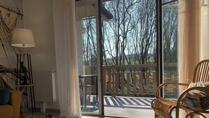 Una sala de estar moderna con una mesa de centro de vidrio, una silla de mimbre y vistas a una terraza de madera y árboles a través de amplias puertas de cristal.