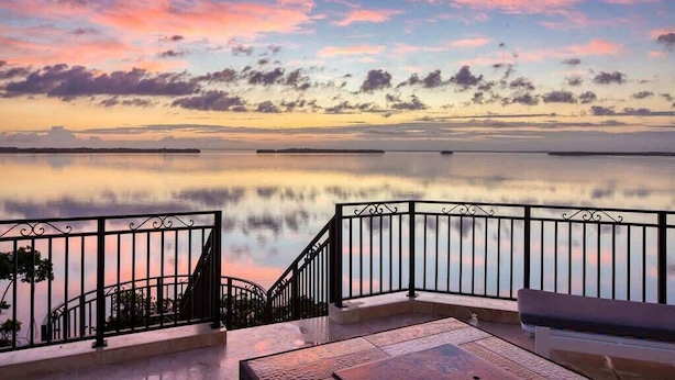 Een balkon met een metalen hekwerk met uitzicht op een kalme waterpartij en een zonsondergang.