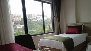 A hotel room with a bed, a table with a red tablecloth, and a window with a view of buildings and trees. - KURT OTEL (Trabzon)