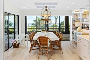 A spacious dining area with a large table and wicker chairs, a modern chandelier, and large windows with a view of palm trees.