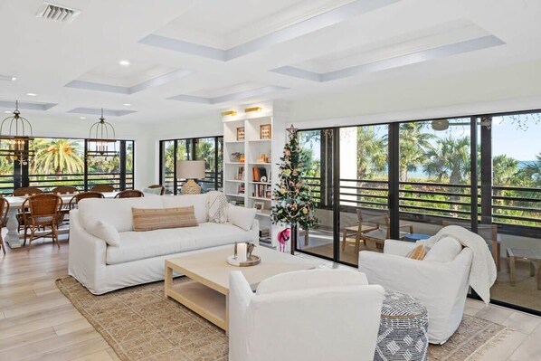 A spacious living area with a white sofa, a coffee table, and a bookshelf. Large windows provide a view of palm trees and a balcony.