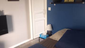 Een slaapkamer met een bed, een nachtkastje met een lamp, een televisie, een lijst met een foto aan de muur en een plank met een bordje met daarop ‘hotdog’.