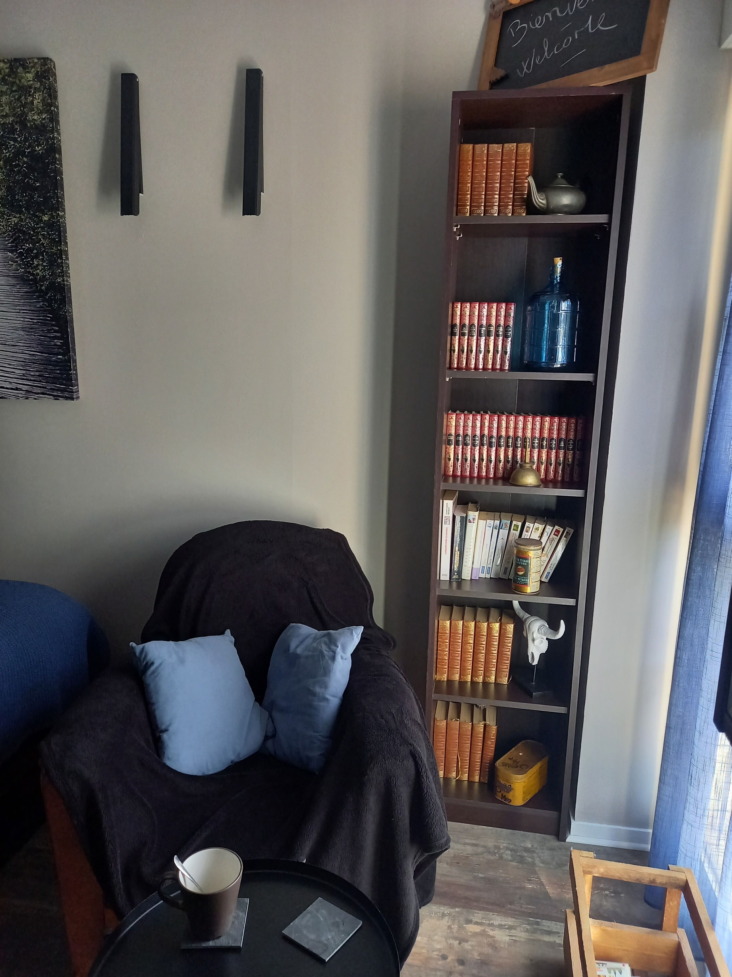 Een gezellig zithoekje met een donkere bank, een klein rond tafeltje met een kop en een boek, en een boekenplank vol boeken en decoratieve objecten.