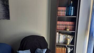 Een gezellig zithoekje met een donkere bank, een klein rond tafeltje met een kop en een boek, en een boekenplank vol boeken en decoratieve objecten.