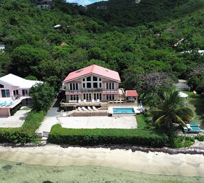 A large house with a red roof, a swimming pool, and a beach in the foreground.