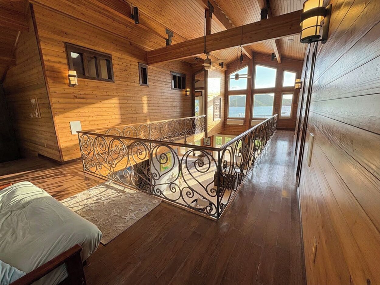 A wooden interior with a bed, a railing with intricate designs, and large windows.