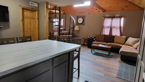A kitchen with a central island, a dining area with chairs, a staircase, a living room with a sofa, and a clock on the wall. - Northwoods Cabin near Upper Red Lake (Kelliher)