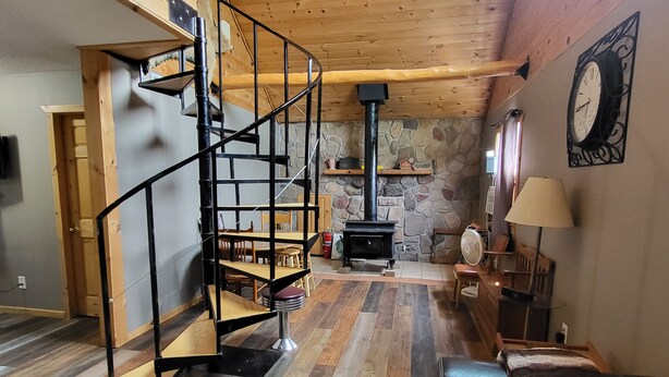 A spacious living area with a wooden staircase, a stone fireplace, and a clock on the wall. - Northwoods Cabin near Upper Red Lake (Kelliher)