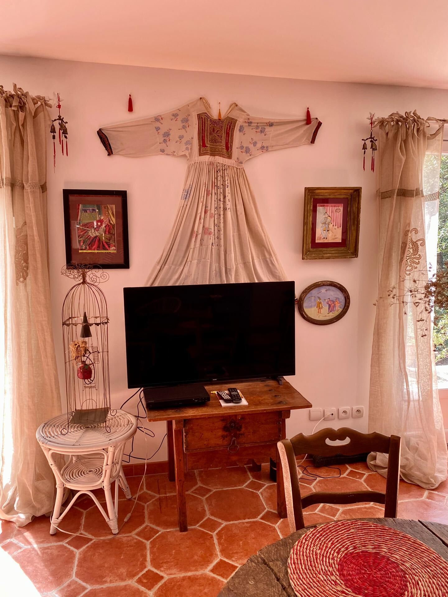 A cozy living room with a large flat-screen TV, a wooden coffee table, a wicker chair, and a decorative dress hanging on the wall.
