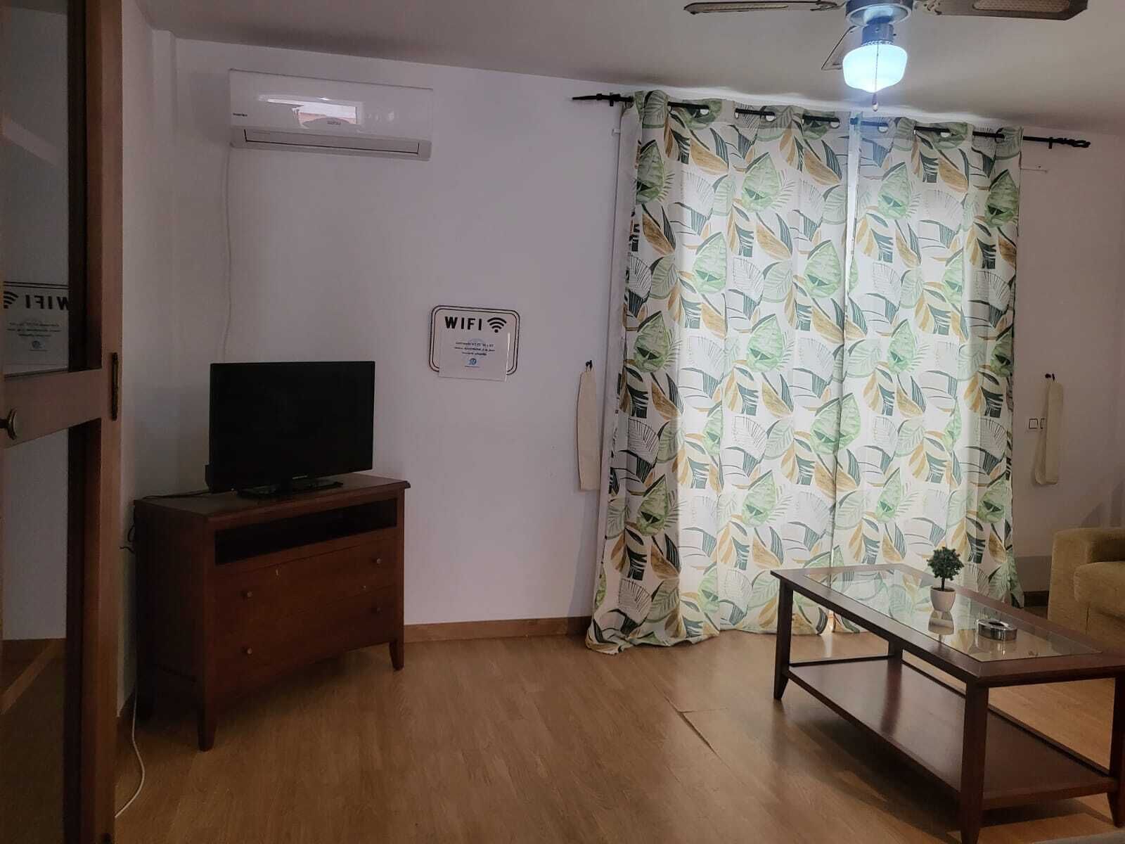 A living room with a flat-screen TV on a wooden cabinet, a glass coffee table, a ceiling fan, and a large window with a leaf-patterned curtain.
