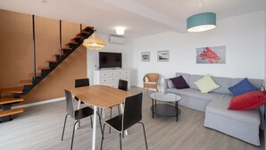 A modern living room with a staircase, a dining area, and a sofa with cushions. - Apartment Duplex T1 - Duplex WEST (Arco da Calheta)