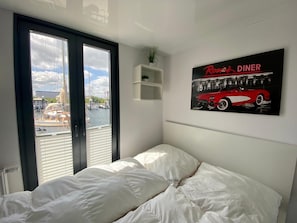 A bedroom with a bed, a window with blinds, a small shelf with a plant, and a poster of a red car on the wall.