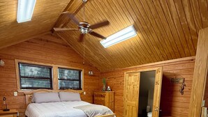 Un dormitorio de cabaña de madera con cama, mesitas de noche, cómoda, ventilador de techo y ventana con persianas.