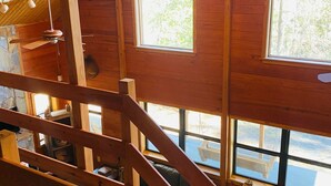 Interior de una cabaña de madera con escalera, ventanas amplias y un candelabro.