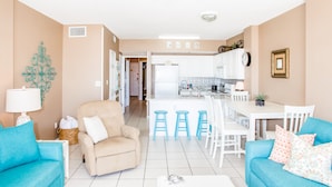 A modern living room with two blue sofas, a beige armchair, and a white coffee table. A kitchen with white cabinets and a dining area with blue stools is visible in the background. - Direct Beach Front! You gotta see this view! (Gulf Shores)