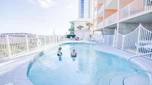 A swimming pool with two people in the water, surrounded by a white fence and a building with balconies. - Direct Beach Front! You gotta see this view! (Gulf Shores)