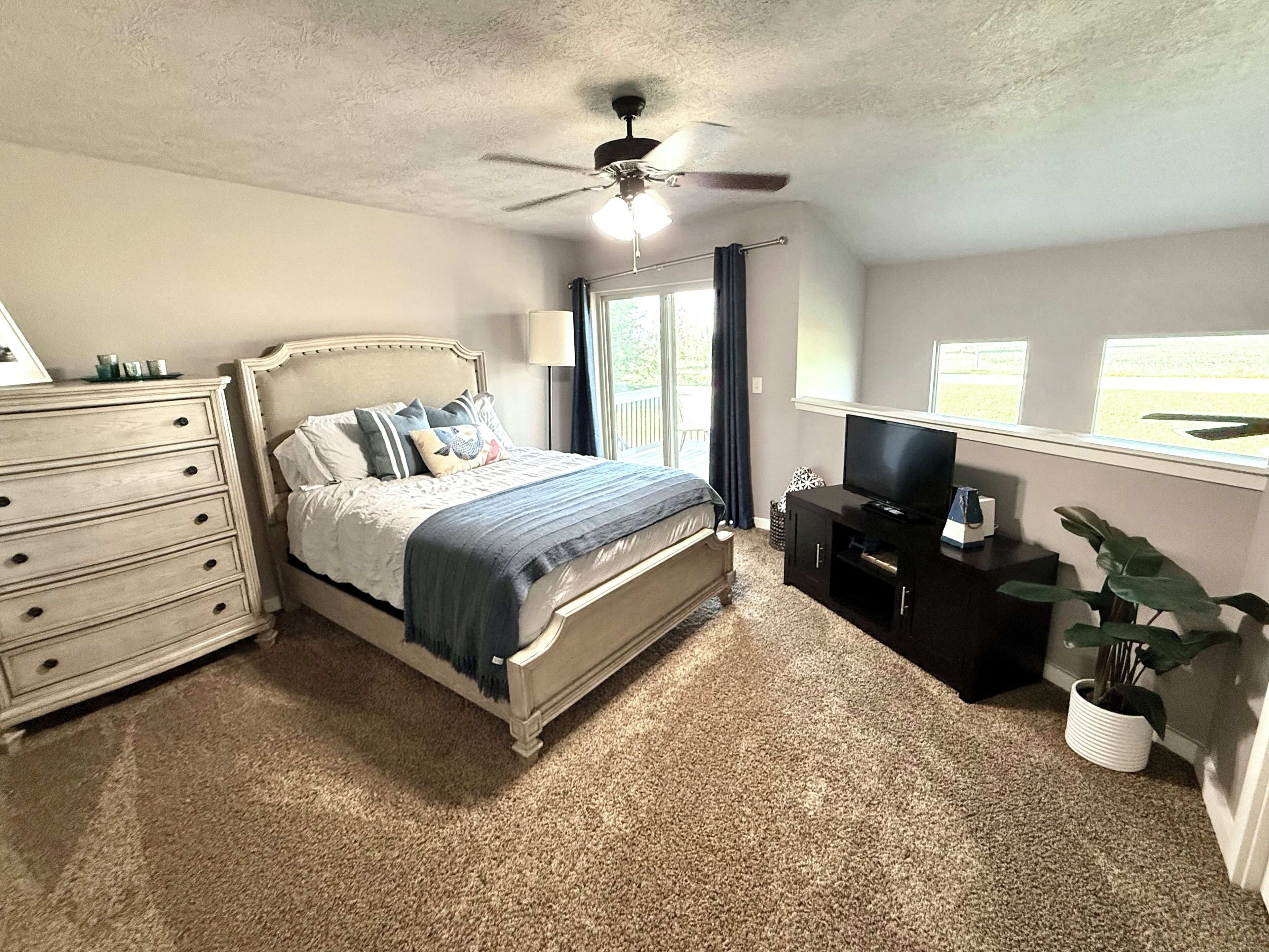 A bedroom with a bed, dresser, TV stand, ceiling fan, and large windows.