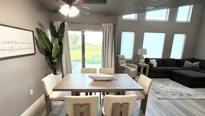 Un espace repas avec une table et des chaises en bois, un ventilateur de plafond et une vue sur l’extérieur grâce à de grandes fenêtres.