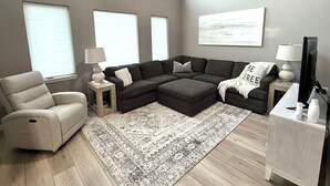 A living room with a sectional sofa, a recliner, a coffee table, a TV, and a large window with blinds.