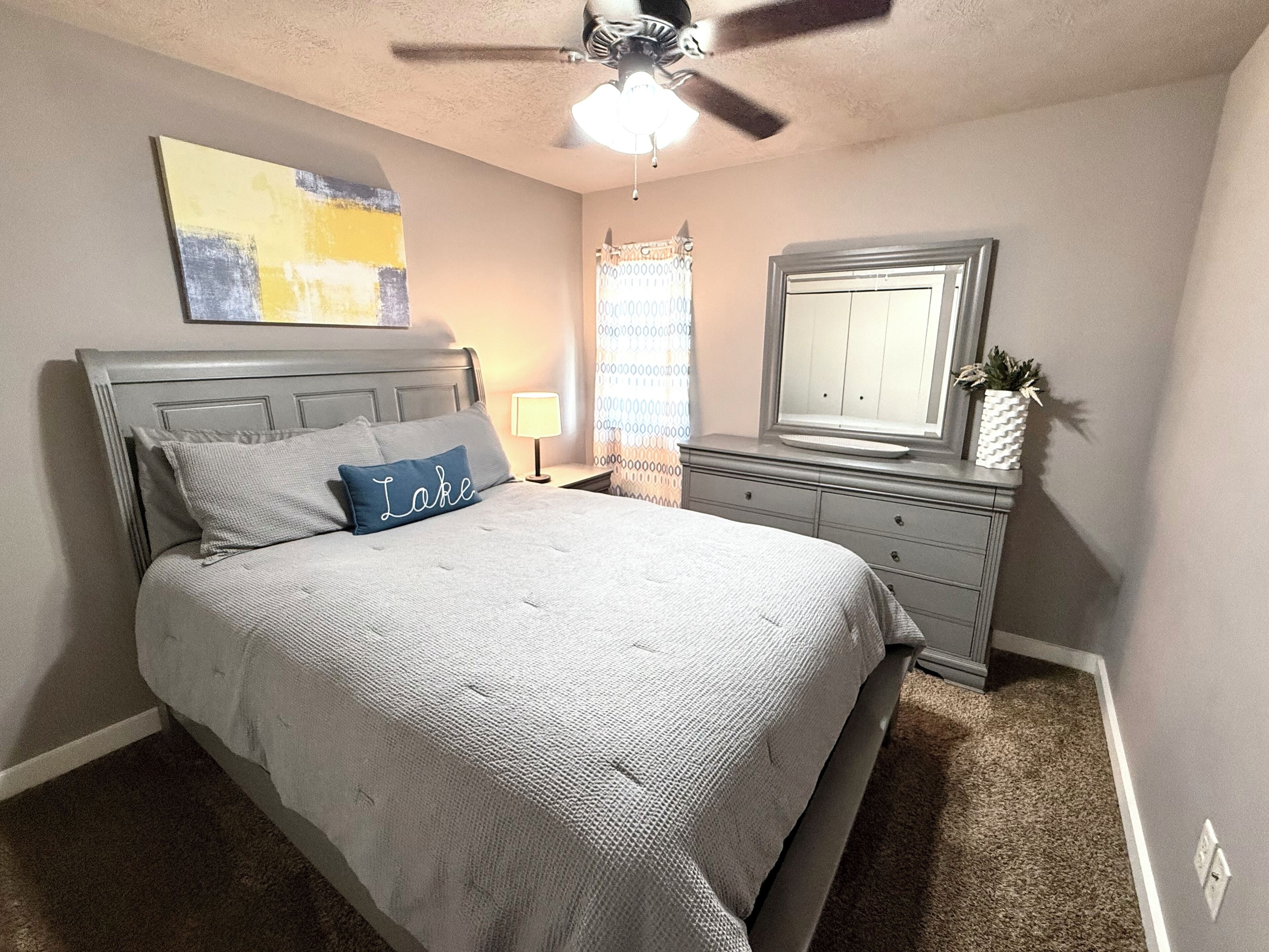 A bedroom with a bed, a dresser, a ceiling fan, a window with curtains, and a painting on the wall.