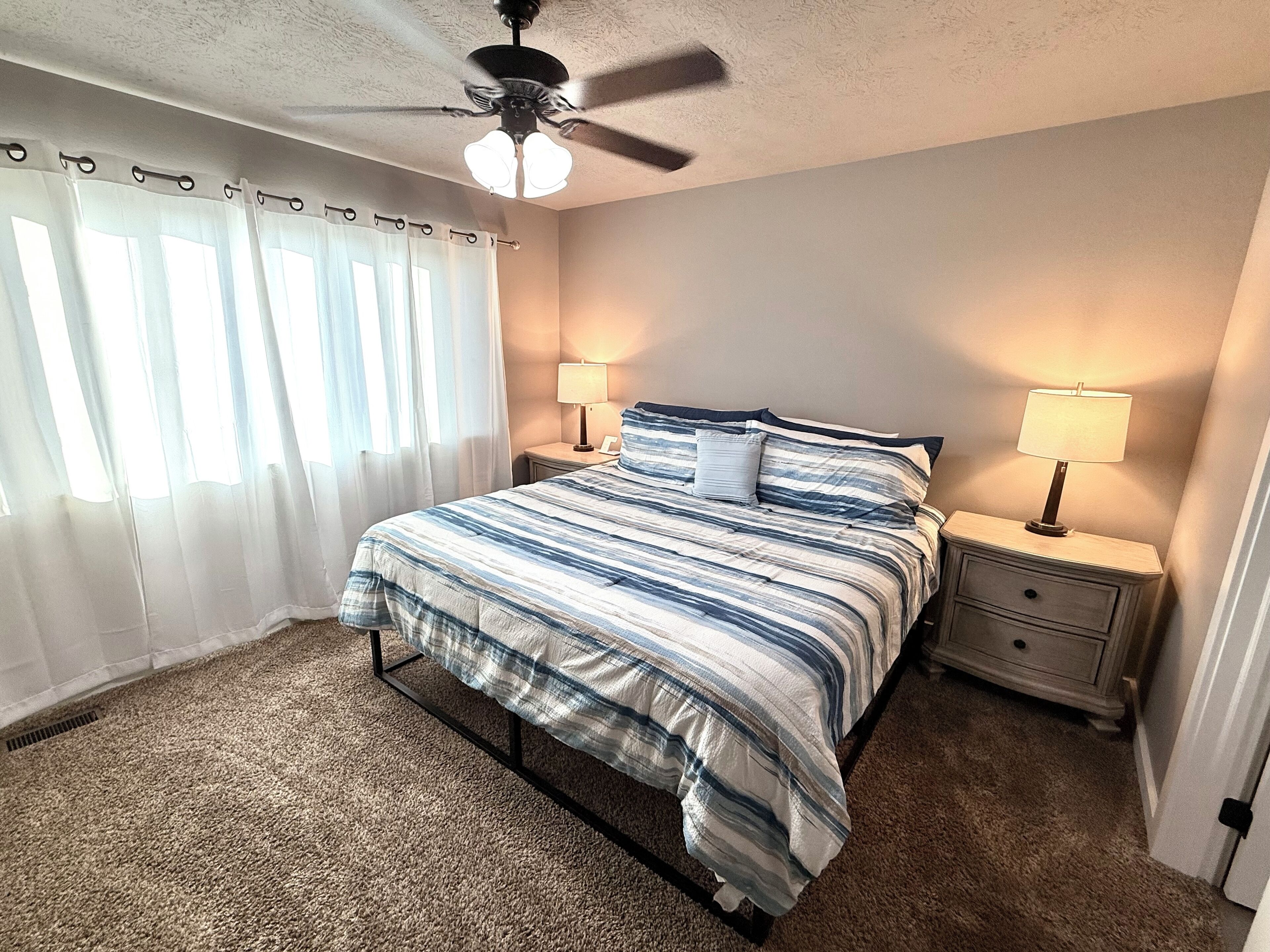 A bedroom with a bed, two bedside tables with lamps, a ceiling fan, and a window with curtains.