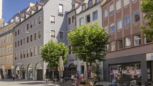Vista de una calle con diversos edificios, incluyendo un edificio blanco destacado con techo triangular y otro marrón con grandes ventanales. Se ven árboles, mesas al aire libre y bicicletas estacionadas a lo largo de la acera.
