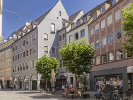 Vista de una calle con diversos edificios, incluyendo un edificio blanco destacado con techo triangular y otro marrón con grandes ventanales. Se ven árboles, mesas al aire libre y bicicletas estacionadas a lo largo de la acera.