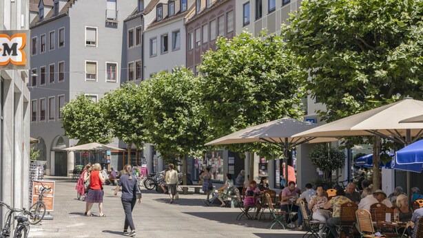 Escena animada de una calle con terrazas de café, peatones y edificios de diversos estilos arquitectónicos.