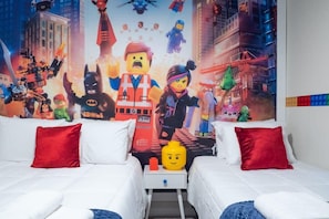 Quarto de hotel com duas camas, um travesseiro vermelho, um travesseiro branco, uma pequena mesa e um mural temático da LEGO.