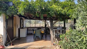 En overdækket terrasse med spisebord og stole, omgivet af frodigt grønt og udsigt til naturen.