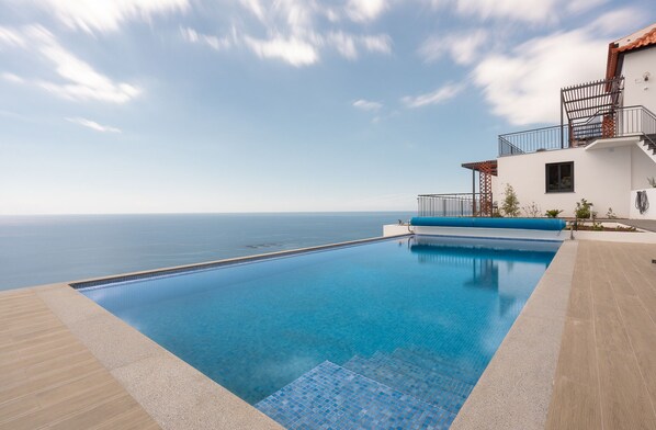 A swimming pool with a view of the sea, a white building with a balcony, and a clear sky.