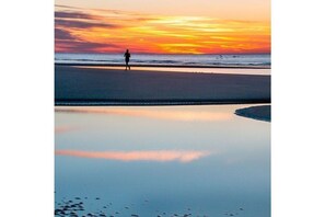 Escena de playa con una persona caminando sobre la arena, agua tranquila que refleja el cielo y un atardecer.