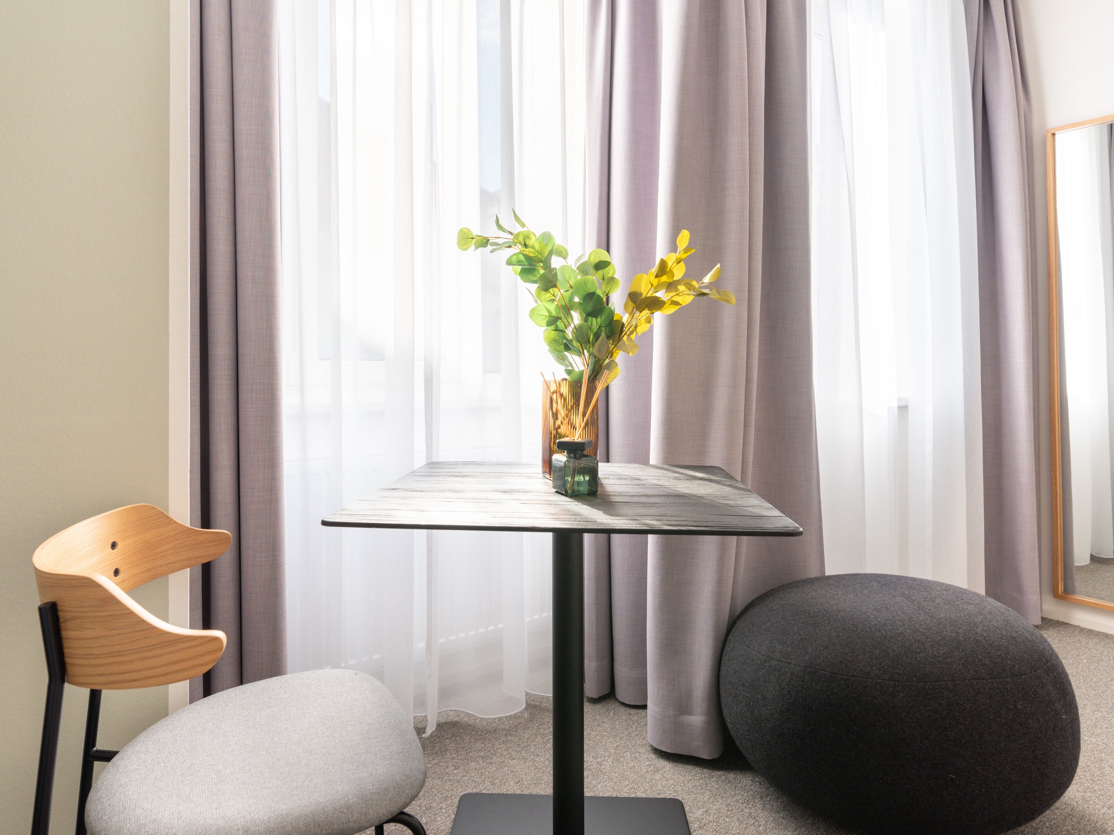 A modern room with a glass table, a wooden chair, a grey cushioned seat, and a black ottoman.