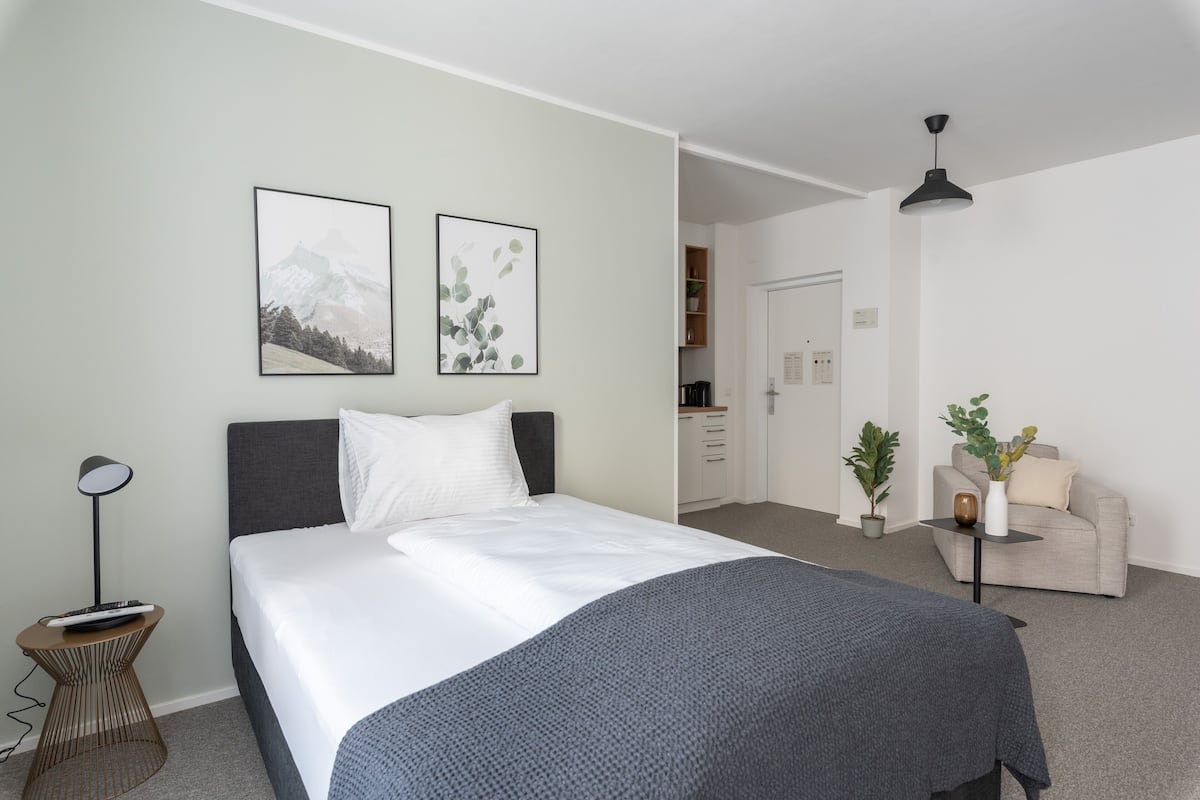 A modern hotel room with a large bed, a bedside table with a lamp, a small side table, a sofa, and a potted plant.