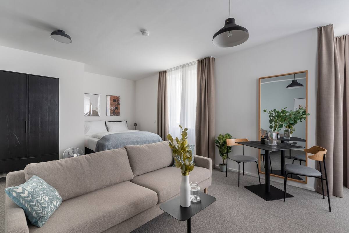 A modern, well-lit living room with a sofa, a dining table with chairs, a large mirror, and a wardrobe.