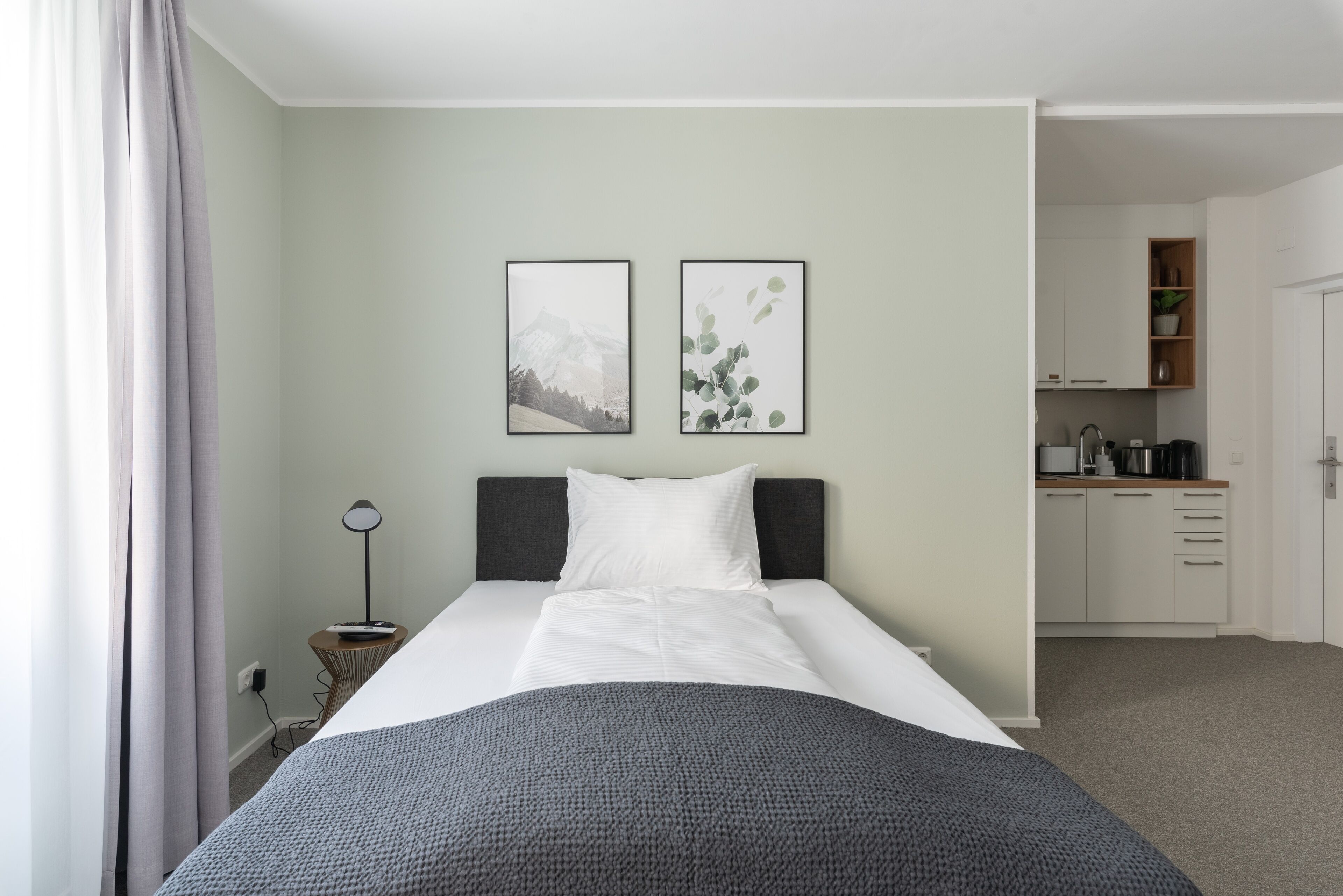 Un dormitorio ordenado con cama, lámpara de noche y vista hacia una zona de cocina.