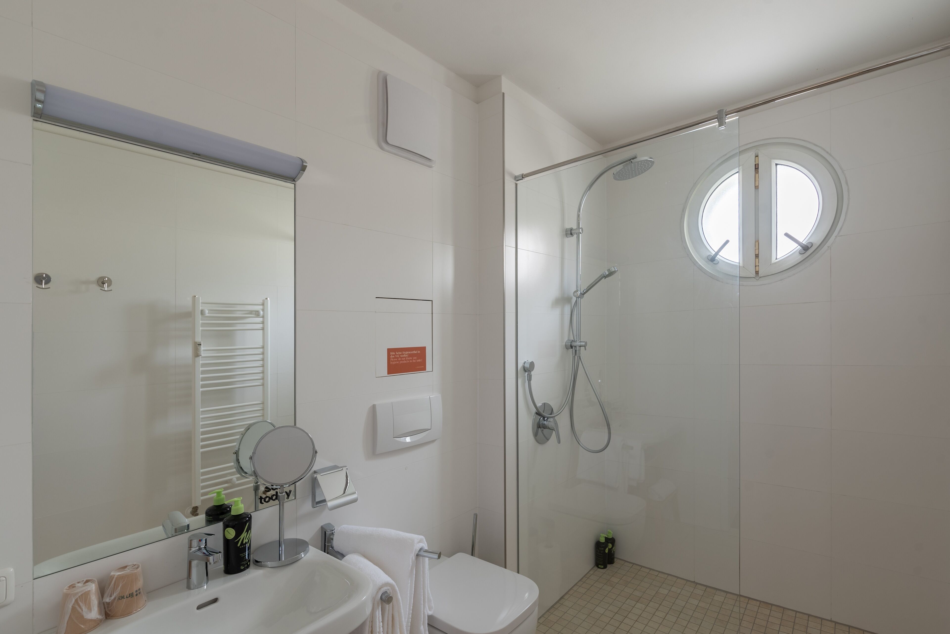 Un baño moderno con cabina de ducha de vidrio, un ventanal redondo y un espejo grande sobre el lavamanos.