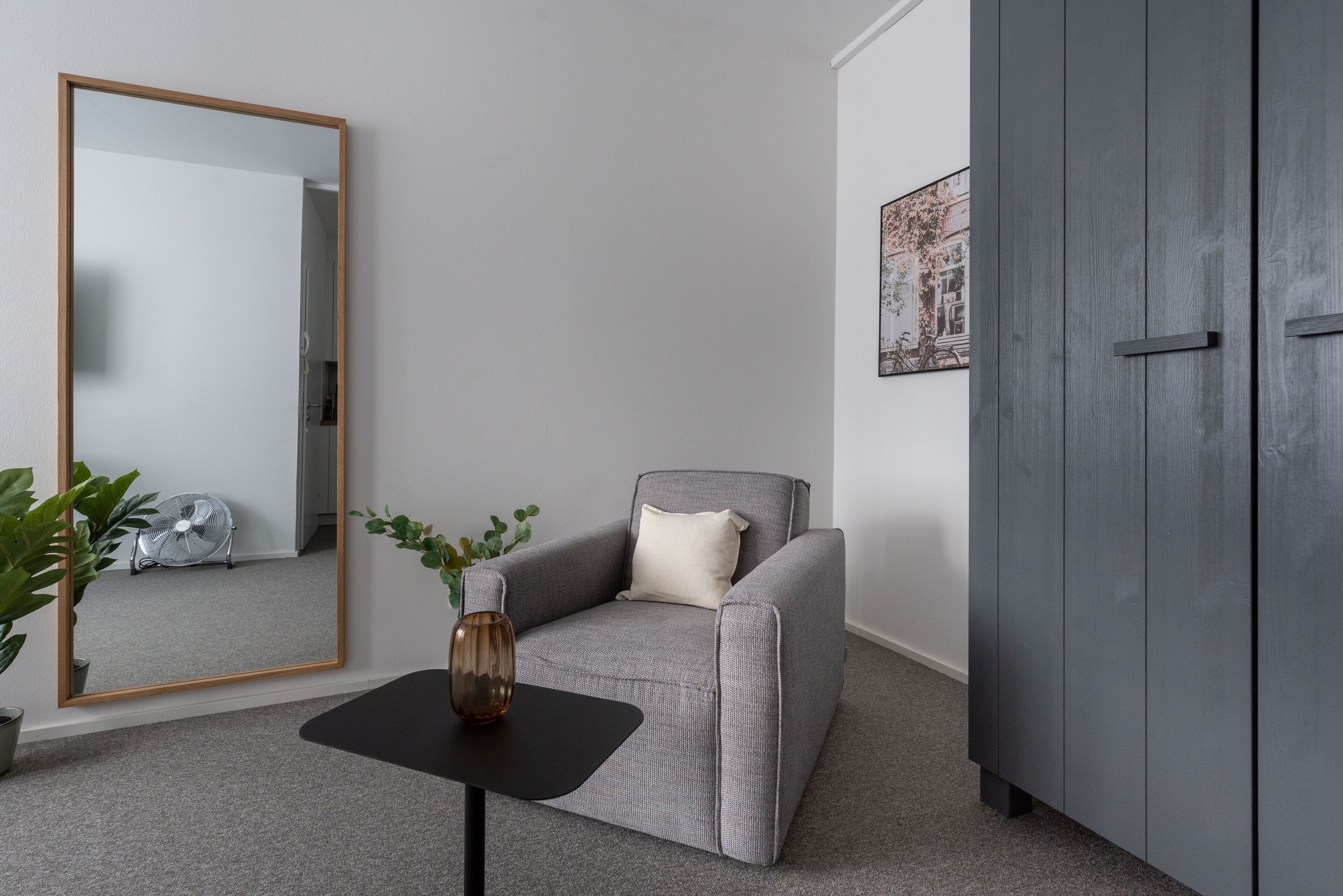 Une pièce comprenant un fauteuil gris, une table d’appoint noire avec un vase, un grand miroir, une armoire grise et un tableau encadré accroché au mur.