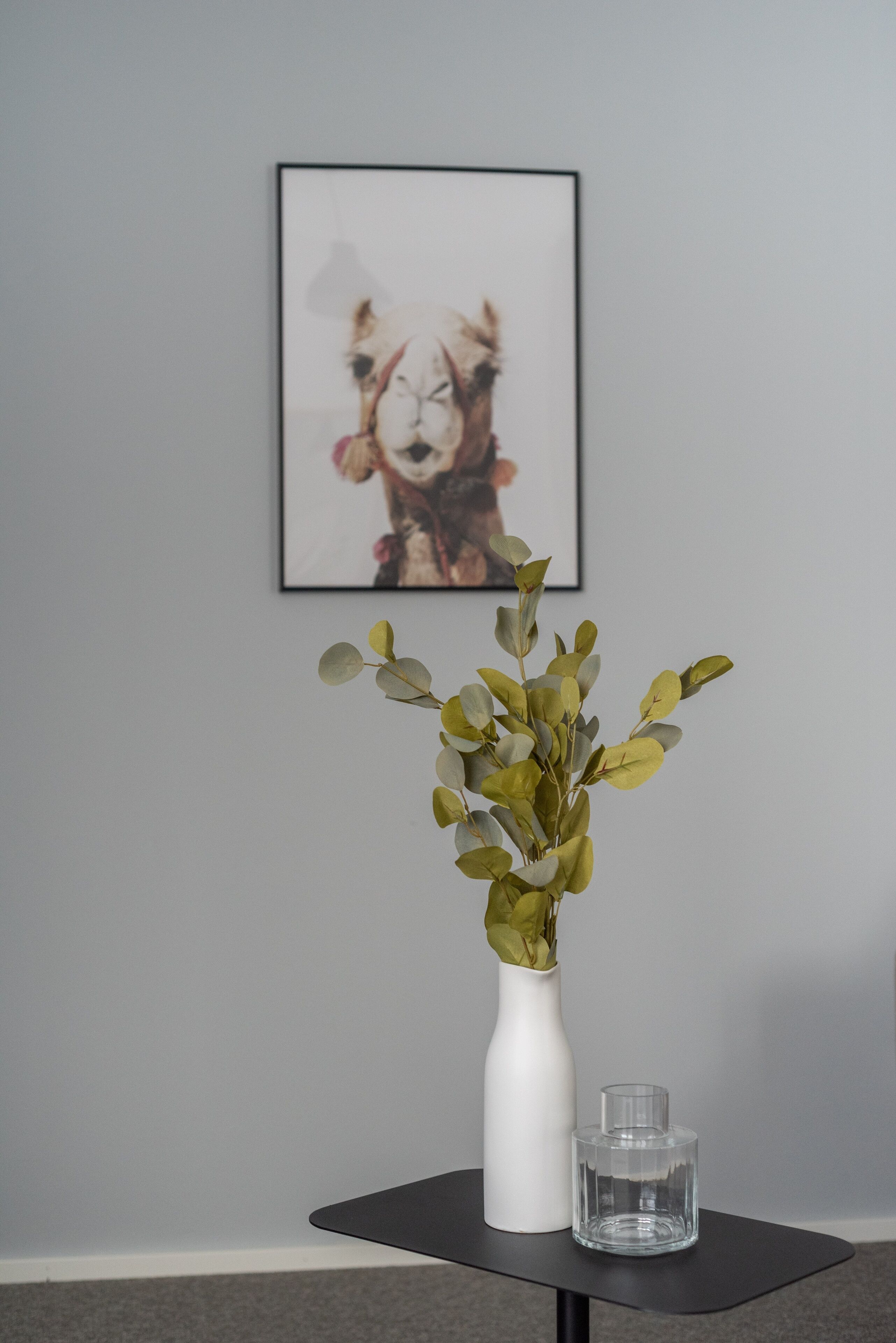 Eine weiße Vase mit grünen Blättern auf einem schwarzen Tisch vor einer grauen Wand, darüber ein gerahmtes Bild.