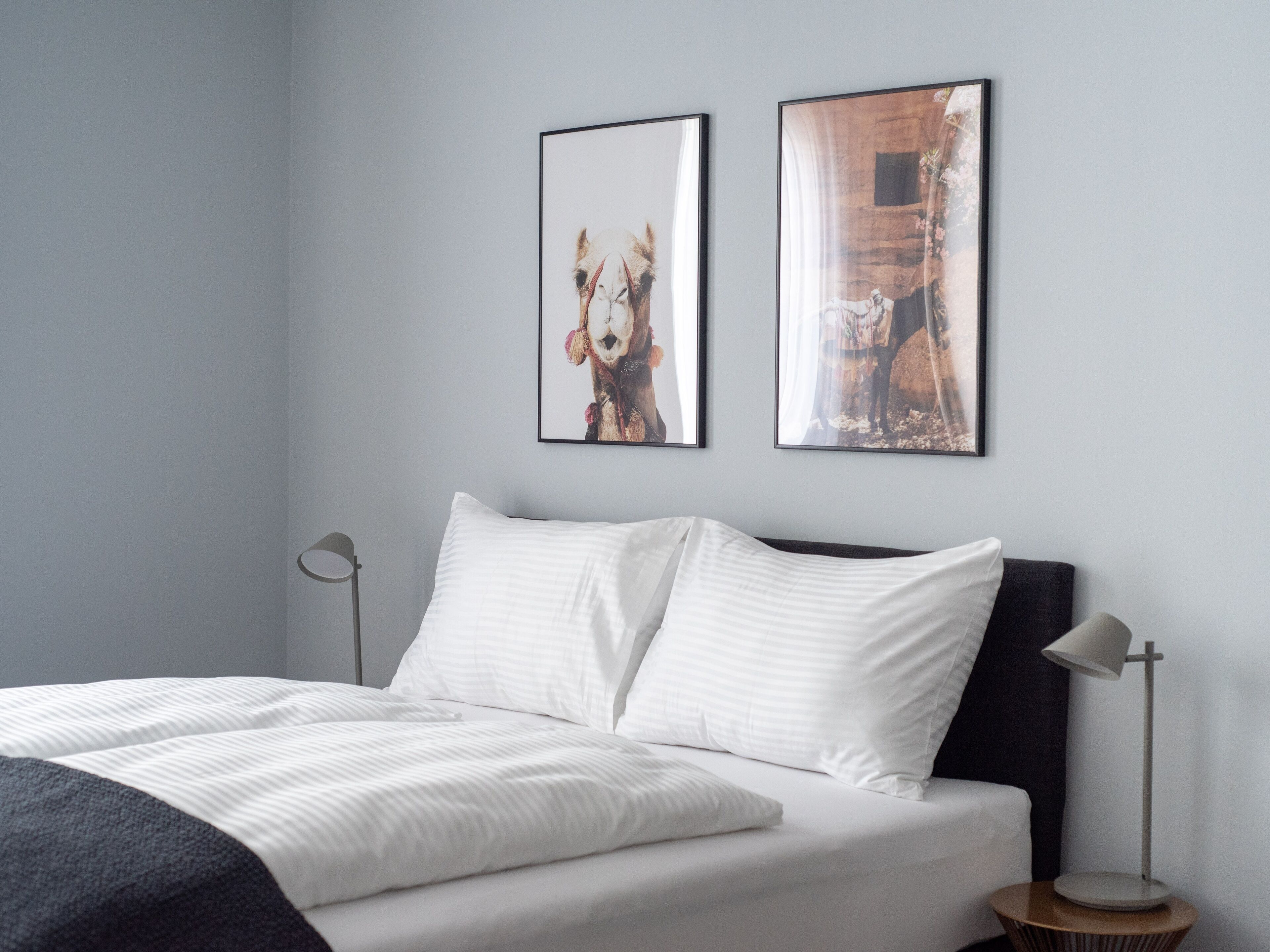 Una cama bien hecha con sábanas blancas y dos almohadas, una mesita de noche con una lámpara y dos cuadros enmarcados en la pared.