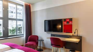 A hotel room with a bed, a desk with a TV, two red chairs, a small table, and a window with curtains.