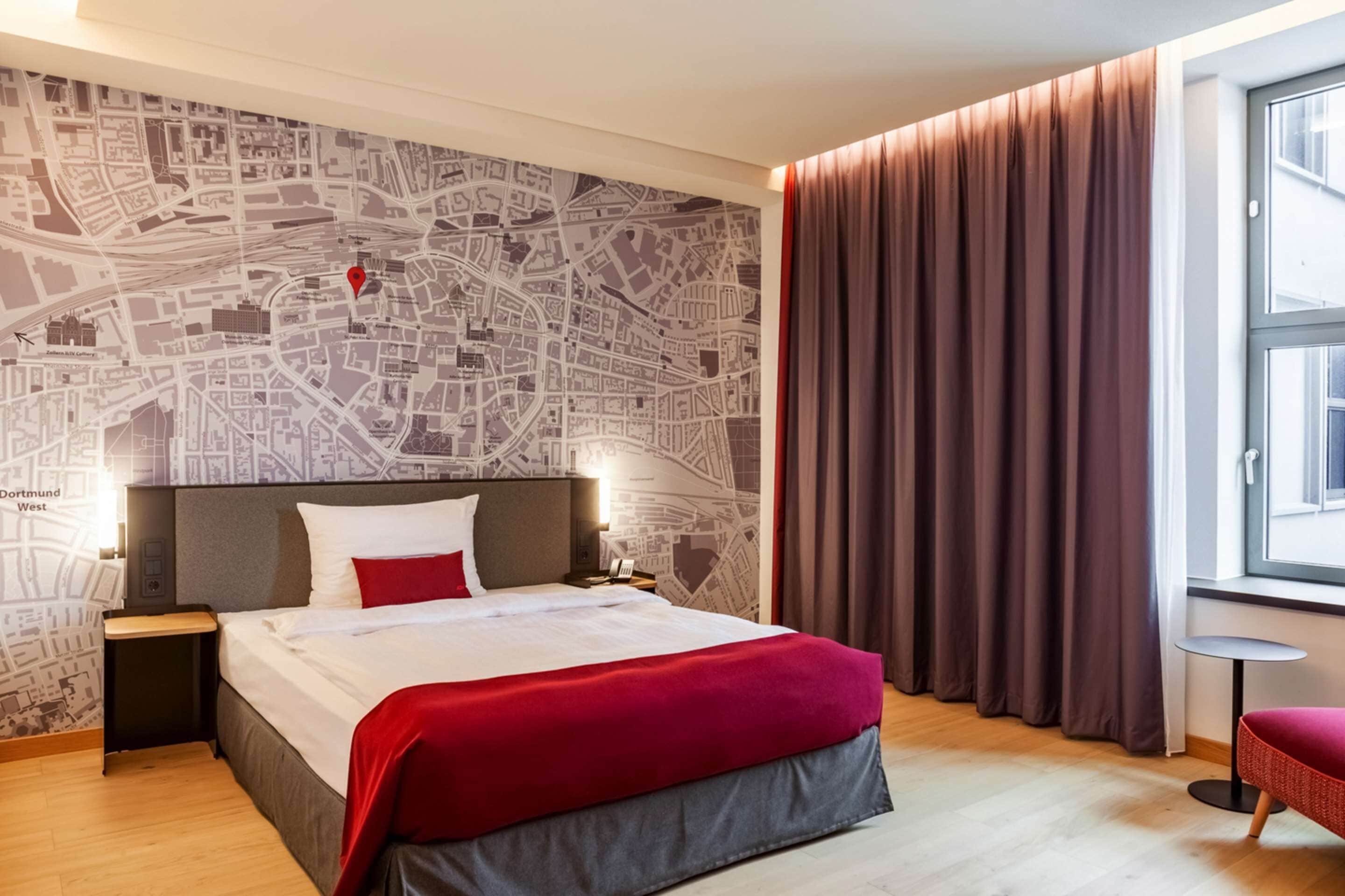 A hotel room with a large bed, a map wall, a window with curtains, and a small table.