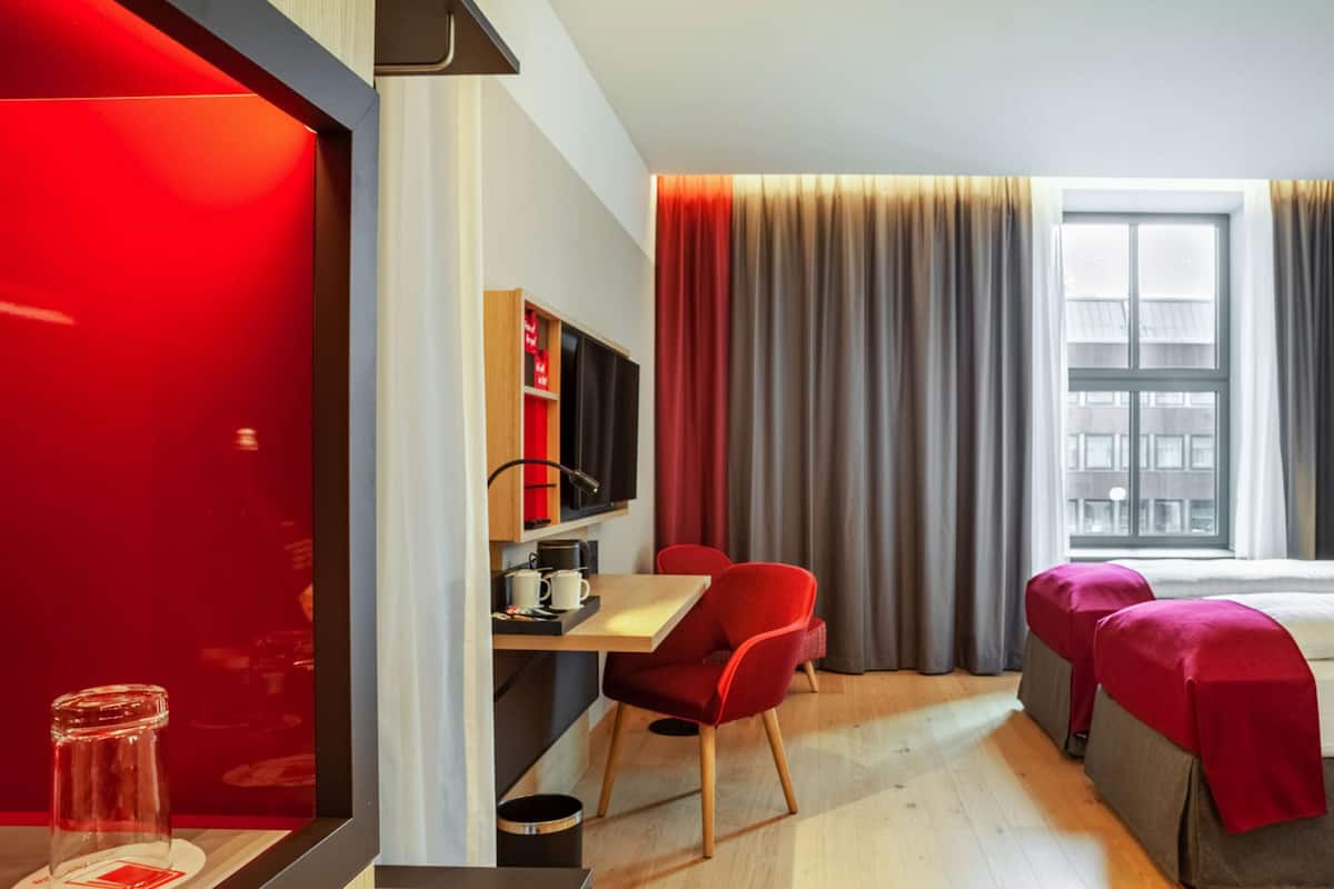 A modern hotel room with a wooden desk, red chairs, a bed with red covers, a bookshelf, and a window with curtains.