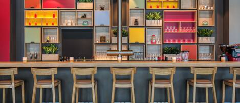 A bar area with wooden stools, shelves with bottles and glasses, and a dark-colored wall.