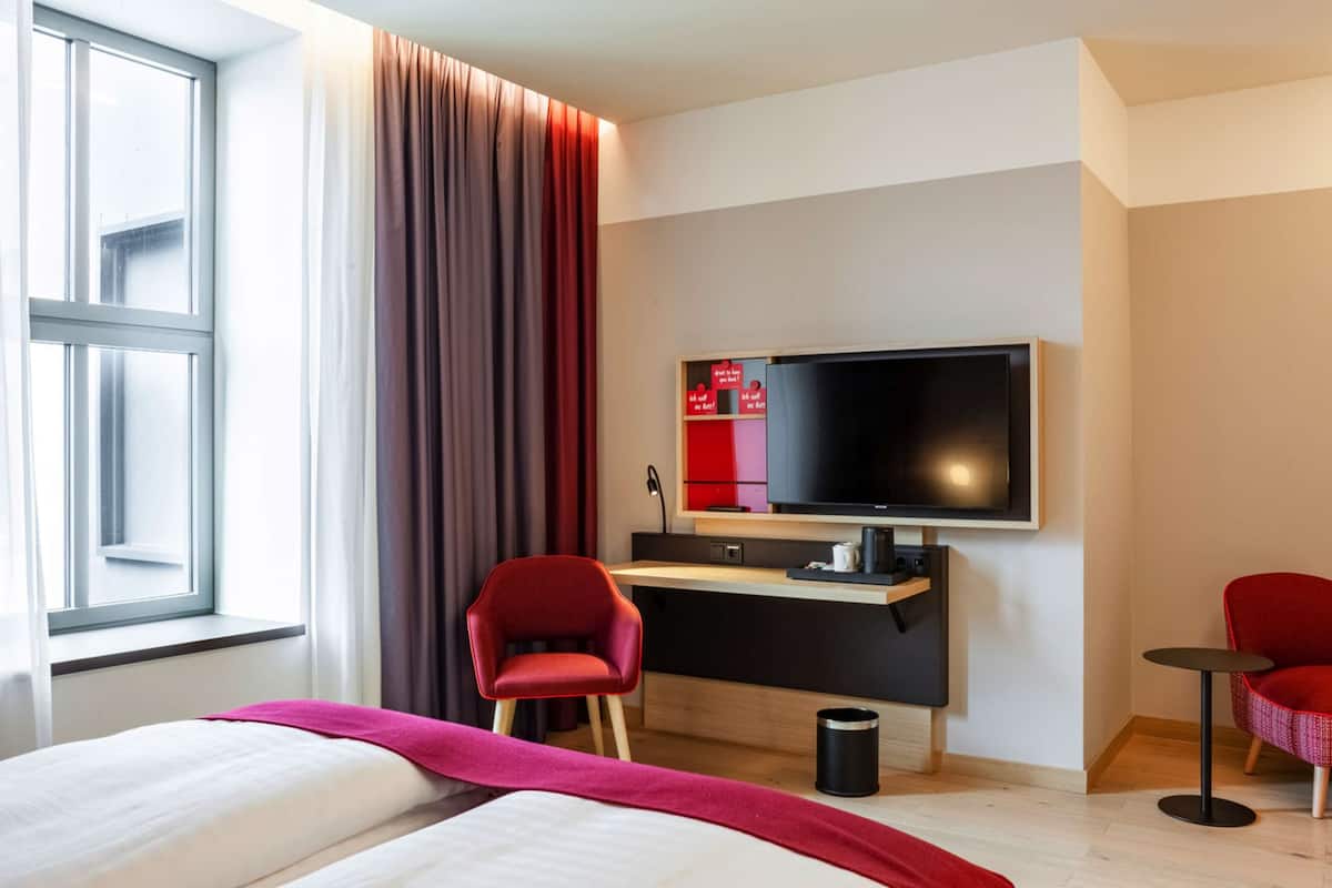 A hotel room with a bed, a desk, a television, and two red chairs.
